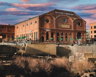 A large brick building with arched windows, displaying signs for REI Co-op and Starbucks Coffee, stands prominently against a backdrop of colorful clouds. There is a patio area with green umbrellas, suggesting an outdoor seating arrangement. The foreground shows a body of water and some dry bushes, adding a natural element to the urban setting.