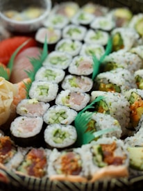 A variety of sushi rolls are arranged in a decorative manner, displaying a range of ingredients such as fish, cucumber, and other colorful vegetables. The rolls are tightly packed with rice and seaweed, showcasing different textures and flavors. The presentation includes vibrant green garnishes that enhance the visual appeal.