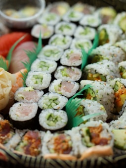 A variety of sushi rolls are arranged in a decorative manner, displaying a range of ingredients such as fish, cucumber, and other colorful vegetables. The rolls are tightly packed with rice and seaweed, showcasing different textures and flavors. The presentation includes vibrant green garnishes that enhance the visual appeal.
