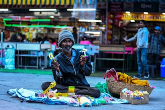 A man sits cross-legged on the ground of a busy market area, holding colorful toys. He wears a beanie and a jacket, and in front of him, there are toys displayed on a cloth spread out. The background shows a bustling market with people and signage illuminated by bright lights.