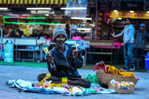 A man sits cross-legged on the ground of a busy market area, holding colorful toys. He wears a beanie and a jacket, and in front of him, there are toys displayed on a cloth spread out. The background shows a bustling market with people and signage illuminated by bright lights.