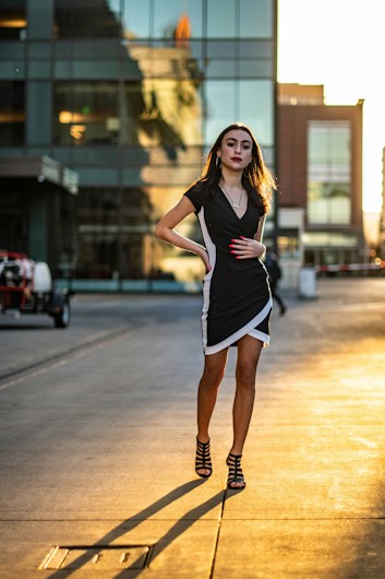 A woman in a black and white dress confidently walks along a sunlit urban street. She has long hair and is wearing black strappy high heels. The background features modern glass buildings reflecting the evening sunlight, creating a warm and stylish atmosphere.