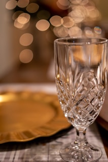 An elegant crystal glass with intricate patterns stands on a table, alongside a gold plate. Soft bokeh lights create a warm, festive ambiance in the background, enhancing the luxurious setting.