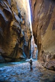 A person wearing outdoor gear stands in a narrow canyon with towering rock walls on either side. The ground is covered with shallow water, and a hiking stick is being used for balance. Sunlight illuminates the upper portions of the canyon, creating a contrast between the light and shadowed areas.