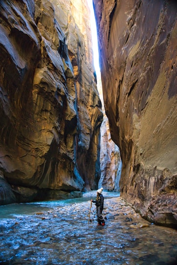 A person wearing outdoor gear stands in a narrow canyon with towering rock walls on either side. The ground is covered with shallow water, and a hiking stick is being used for balance. Sunlight illuminates the upper portions of the canyon, creating a contrast between the light and shadowed areas.