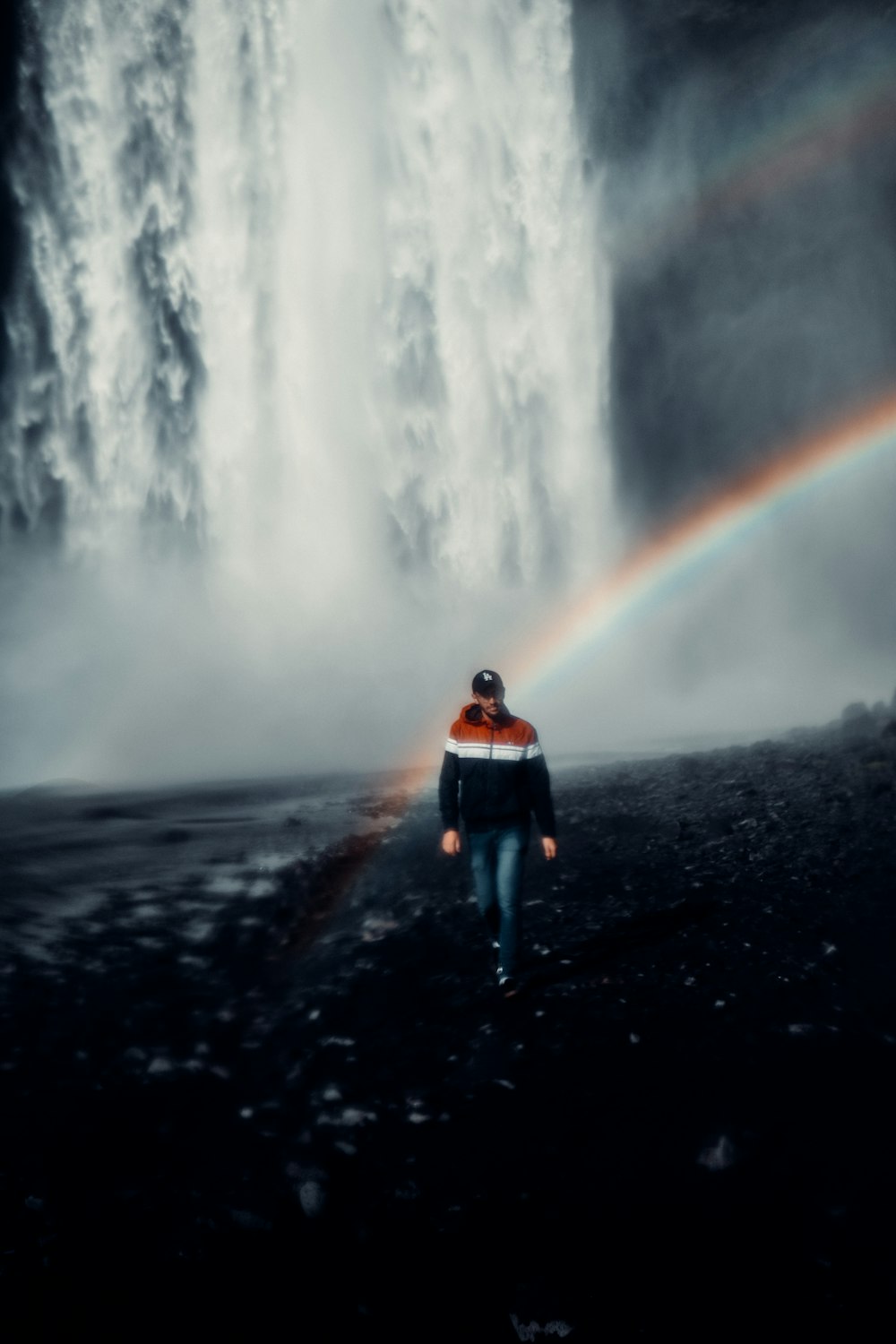 man in orange jacket and blue denim jeans standing on black sand near waterfalls during daytime