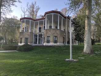 A two-story building with classical architecture, featuring tall white columns and a red-tiled roof. The building is surrounded by lush green grass and several tall trees. The facade is made of beige bricks, with large, dark-framed windows.