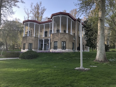 A two-story building with classical architecture, featuring tall white columns and a red-tiled roof. The building is surrounded by lush green grass and several tall trees. The facade is made of beige bricks, with large, dark-framed windows.