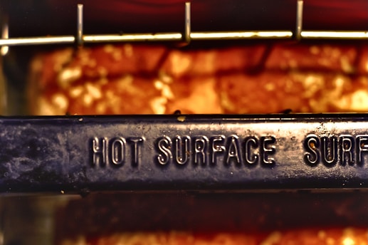 A metal surface with the words 'HOT SURFACE' embossed on it, set against a blurred background with warm, glowing colors. The surface appears to be part of a cooking or heating appliance.
