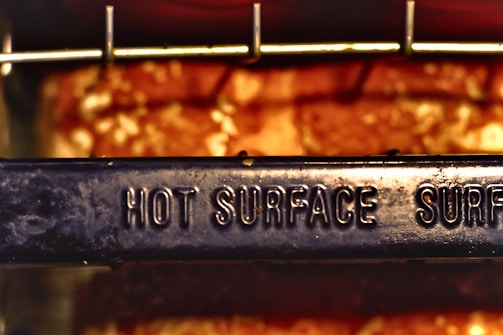 A metal surface with the words 'HOT SURFACE' embossed on it, set against a blurred background with warm, glowing colors. The surface appears to be part of a cooking or heating appliance.