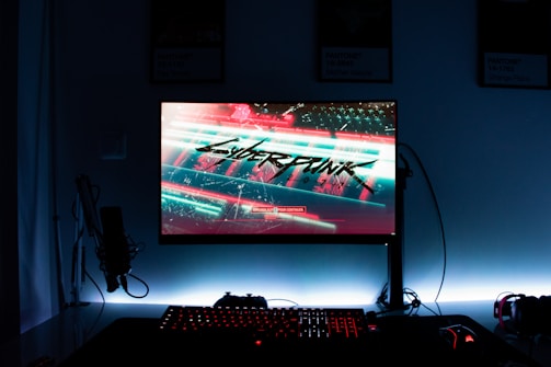 A gaming setup features a glowing monitor displaying a futuristic cityscape with the word 'Cyberpunk' prominently featured on the screen. The setup includes a backlit keyboard and a computer mouse, with a microphone and headphones to the side. The ambiance is dark with illuminated accents.