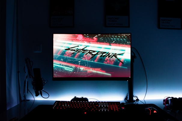 A gaming setup features a glowing monitor displaying a futuristic cityscape with the word 'Cyberpunk' prominently featured on the screen. The setup includes a backlit keyboard and a computer mouse, with a microphone and headphones to the side. The ambiance is dark with illuminated accents.