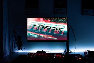 A gaming setup features a glowing monitor displaying a futuristic cityscape with the word 'Cyberpunk' prominently featured on the screen. The setup includes a backlit keyboard and a computer mouse, with a microphone and headphones to the side. The ambiance is dark with illuminated accents.