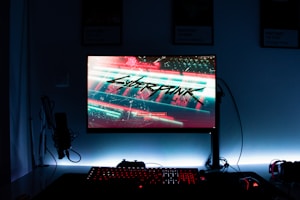 A gaming setup features a glowing monitor displaying a futuristic cityscape with the word 'Cyberpunk' prominently featured on the screen. The setup includes a backlit keyboard and a computer mouse, with a microphone and headphones to the side. The ambiance is dark with illuminated accents.