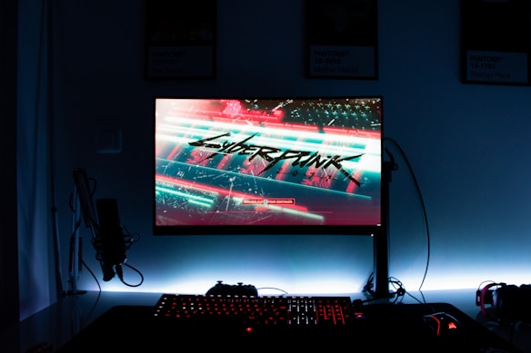 A gaming setup features a glowing monitor displaying a futuristic cityscape with the word 'Cyberpunk' prominently featured on the screen. The setup includes a backlit keyboard and a computer mouse, with a microphone and headphones to the side. The ambiance is dark with illuminated accents.
