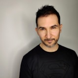 A man with short dark hair and a neatly trimmed beard is wearing a black shirt with a Superman logo. The background is a plain light grey, and he has a slight smile with a direct gaze.