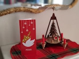 A red holiday card with snowflakes and a white dove symbol alongside a wooden Christmas pyramid featuring small figures and red candles. Both items rest on a red plaid cloth.