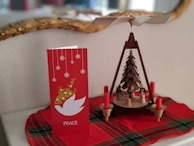 A red holiday card with snowflakes and a white dove symbol alongside a wooden Christmas pyramid featuring small figures and red candles. Both items rest on a red plaid cloth.