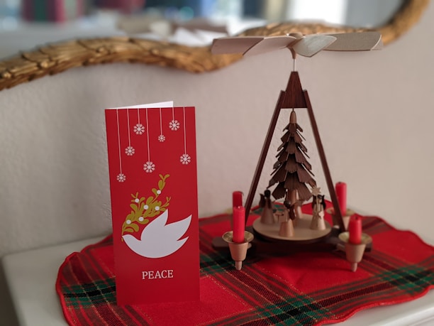 A red holiday card with snowflakes and a white dove symbol alongside a wooden Christmas pyramid featuring small figures and red candles. Both items rest on a red plaid cloth.