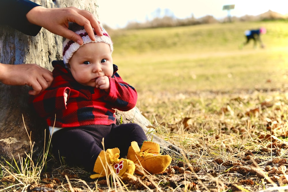 child in red and black jacket sitting on dried leaves on ground during daytime