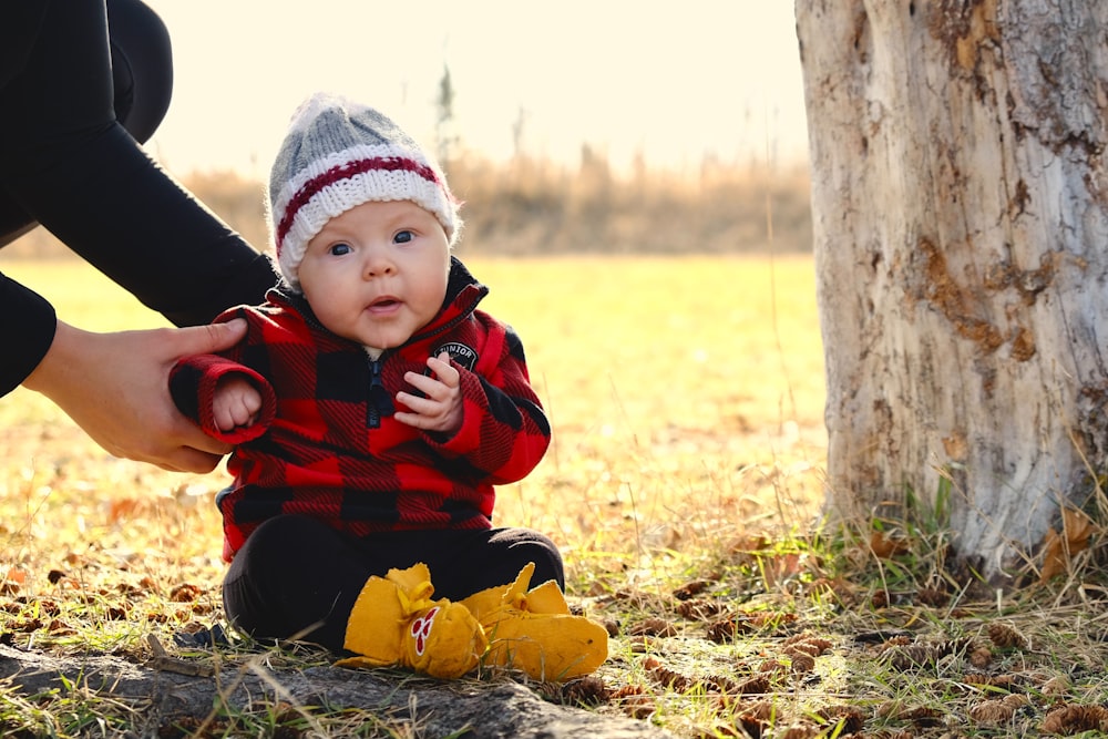 child in black and red jacket sitting on ground beside tree during daytime