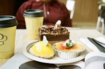 A table with an assortment of desserts including a chocolate cake with chocolate shavings, a yellow-glazed dessert with coconut flakes, and a creme brulee topped with a sliced strawberry. There are also two takeaway coffee cups. A person in casual clothing is partially visible in the background.