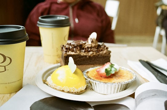 A table with an assortment of desserts including a chocolate cake with chocolate shavings, a yellow-glazed dessert with coconut flakes, and a creme brulee topped with a sliced strawberry. There are also two takeaway coffee cups. A person in casual clothing is partially visible in the background.