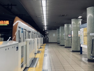 A subway train is at a station platform with vertical pillars and overhead lighting. The train is partially visible and appears to be in motion. The platform features safety barriers and yellow tactile paving. The walls and pillars have a speckled design and there are various signs and maps on display.