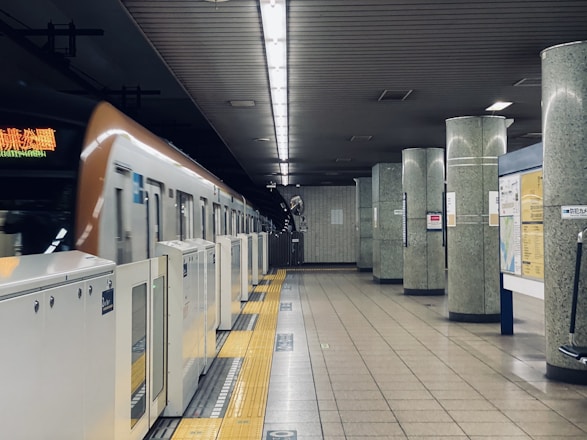 A subway train is at a station platform with vertical pillars and overhead lighting. The train is partially visible and appears to be in motion. The platform features safety barriers and yellow tactile paving. The walls and pillars have a speckled design and there are various signs and maps on display.