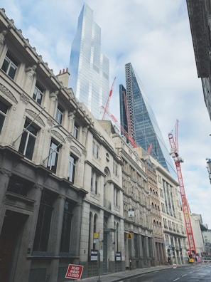 A city street featuring both historic and modern architecture with skyscrapers rising above. Red construction cranes are positioned among the buildings, indicating ongoing development. The sky is mostly cloudy, adding a soft light to the scene.