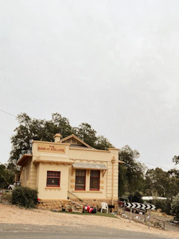 A quaint, historic building labeled 'The Bank of Adelaide' with a yellow and red color scheme, surrounded by trees. The building features large red-framed windows and a neatly maintained front yard with scattered outdoor chairs and a barrel. A directional road sign is visible nearby.