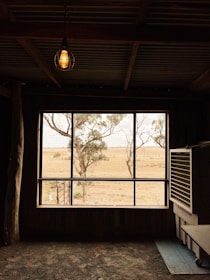 An indoor space with a large window framing a view of an open countryside. The scene outside the window includes dry grass and a few sparse trees. Inside, there is a dimly lit room with a light fixture hanging overhead and an air conditioning unit on the right.