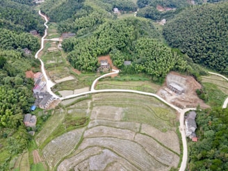 An aerial view of a rural landscape with lush green hills and dense bamboo forests. There are several winding roads leading to scattered houses and small buildings. The image also includes sections of terraced fields and some construction activity visible on the right-hand side.