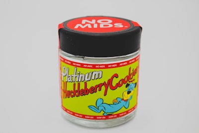 A glass jar with a black lid displaying a colorful label featuring playful cartoon graphics. The label reads 'Platinum Huckleberry Cookies' with a cartoon dog holding a cookie, set against a lime green background. The lid has a bold red label with the words 'NO MIDS' repeated around it.
