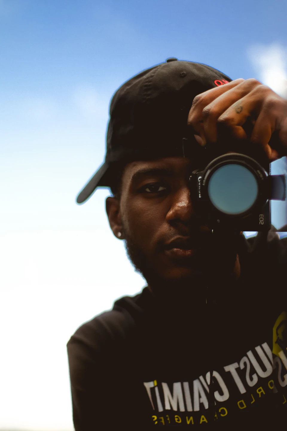 Young man takes photo with camera, wearing black baseball cap and "timimia" t-shirt outdoors