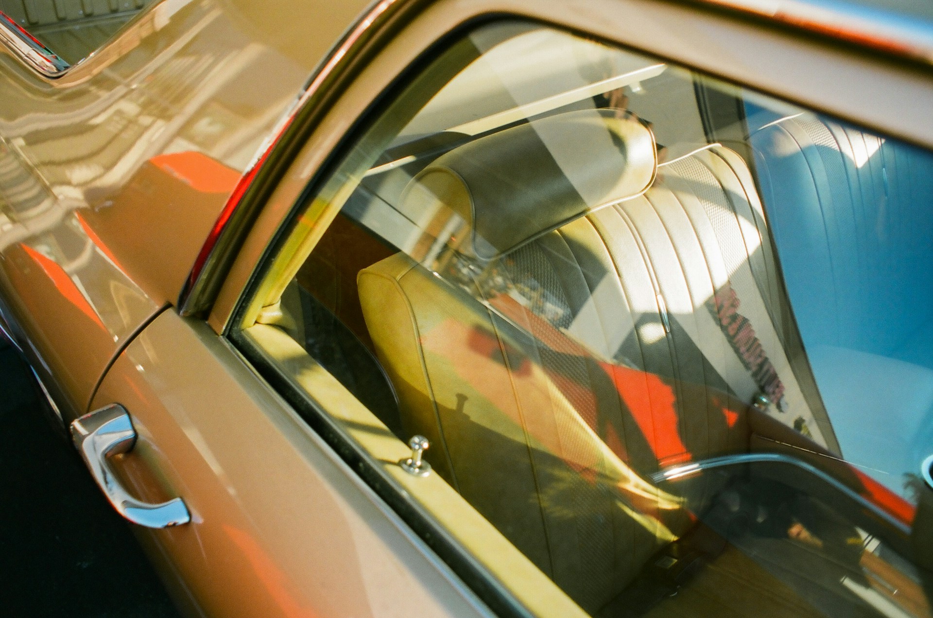 The image captures the interior of a vintage car viewed through the side window. The upholstery is a light tan or beige leather with noticeable stitching. The reflection of the surrounding environment creates interesting patterns on the glass, adding depth to the image.