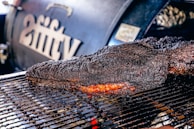 A large, well-cooked piece of meat rests on a grill, displaying a dark, charred crust with a hint of reddish-brown from the smoked interior. In the background, a smoker with branding or lettering is slightly out of focus.