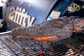 A large, well-cooked piece of meat rests on a grill, displaying a dark, charred crust with a hint of reddish-brown from the smoked interior. In the background, a smoker with branding or lettering is slightly out of focus.