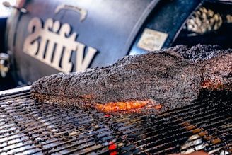 A large, well-cooked piece of meat rests on a grill, displaying a dark, charred crust with a hint of reddish-brown from the smoked interior. In the background, a smoker with branding or lettering is slightly out of focus.