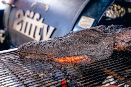 A large, well-cooked piece of meat rests on a grill, displaying a dark, charred crust with a hint of reddish-brown from the smoked interior. In the background, a smoker with branding or lettering is slightly out of focus.