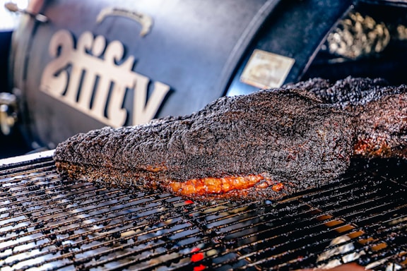 A large, well-cooked piece of meat rests on a grill, displaying a dark, charred crust with a hint of reddish-brown from the smoked interior. In the background, a smoker with branding or lettering is slightly out of focus.