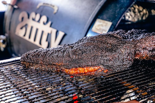 A large, well-cooked piece of meat rests on a grill, displaying a dark, charred crust with a hint of reddish-brown from the smoked interior. In the background, a smoker with branding or lettering is slightly out of focus.
