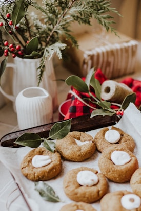 A rustic scene featuring a tray of freshly baked cookies topped with marshmallows, surrounded by green leaves. In the background, a white pitcher holds festive greenery and red berries. A wrapped gift and a spool of twine add to the cozy, holiday atmosphere.