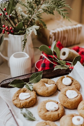 A rustic scene featuring a tray of freshly baked cookies topped with marshmallows, surrounded by green leaves. In the background, a white pitcher holds festive greenery and red berries. A wrapped gift and a spool of twine add to the cozy, holiday atmosphere.