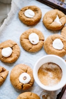 A batch of freshly baked cookies arranged on a baking sheet lined with parchment paper. Each cookie has a soft, round shape and is topped with a small, partially melted marshmallow. The cookies have a golden-brown color, suggesting they are ginger or molasses-based. A white bowl filled with cinnamon sugar is placed among the cookies, adding to the cozy baking setting.