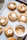 A batch of freshly baked cookies arranged on a baking sheet lined with parchment paper. Each cookie has a soft, round shape and is topped with a small, partially melted marshmallow. The cookies have a golden-brown color, suggesting they are ginger or molasses-based. A white bowl filled with cinnamon sugar is placed among the cookies, adding to the cozy baking setting.