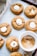 A batch of freshly baked cookies arranged on a baking sheet lined with parchment paper. Each cookie has a soft, round shape and is topped with a small, partially melted marshmallow. The cookies have a golden-brown color, suggesting they are ginger or molasses-based. A white bowl filled with cinnamon sugar is placed among the cookies, adding to the cozy baking setting.