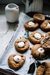 A tray lined with parchment paper holds freshly baked cookies. Each cookie features a distinct pattern and is topped with a small, white marshmallow. Nearby, an empty, white ceramic jug adds a touch of simplicity to the scene. Crumbs are lightly scattered around, emphasizing the fresh, homemade feel. A couple of green leaves are placed on the tray, adding a natural element to the display.
