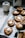 A tray lined with parchment paper holds freshly baked cookies. Each cookie features a distinct pattern and is topped with a small, white marshmallow. Nearby, an empty, white ceramic jug adds a touch of simplicity to the scene. Crumbs are lightly scattered around, emphasizing the fresh, homemade feel. A couple of green leaves are placed on the tray, adding a natural element to the display.