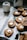 A tray lined with parchment paper holds freshly baked cookies. Each cookie features a distinct pattern and is topped with a small, white marshmallow. Nearby, an empty, white ceramic jug adds a touch of simplicity to the scene. Crumbs are lightly scattered around, emphasizing the fresh, homemade feel. A couple of green leaves are placed on the tray, adding a natural element to the display.